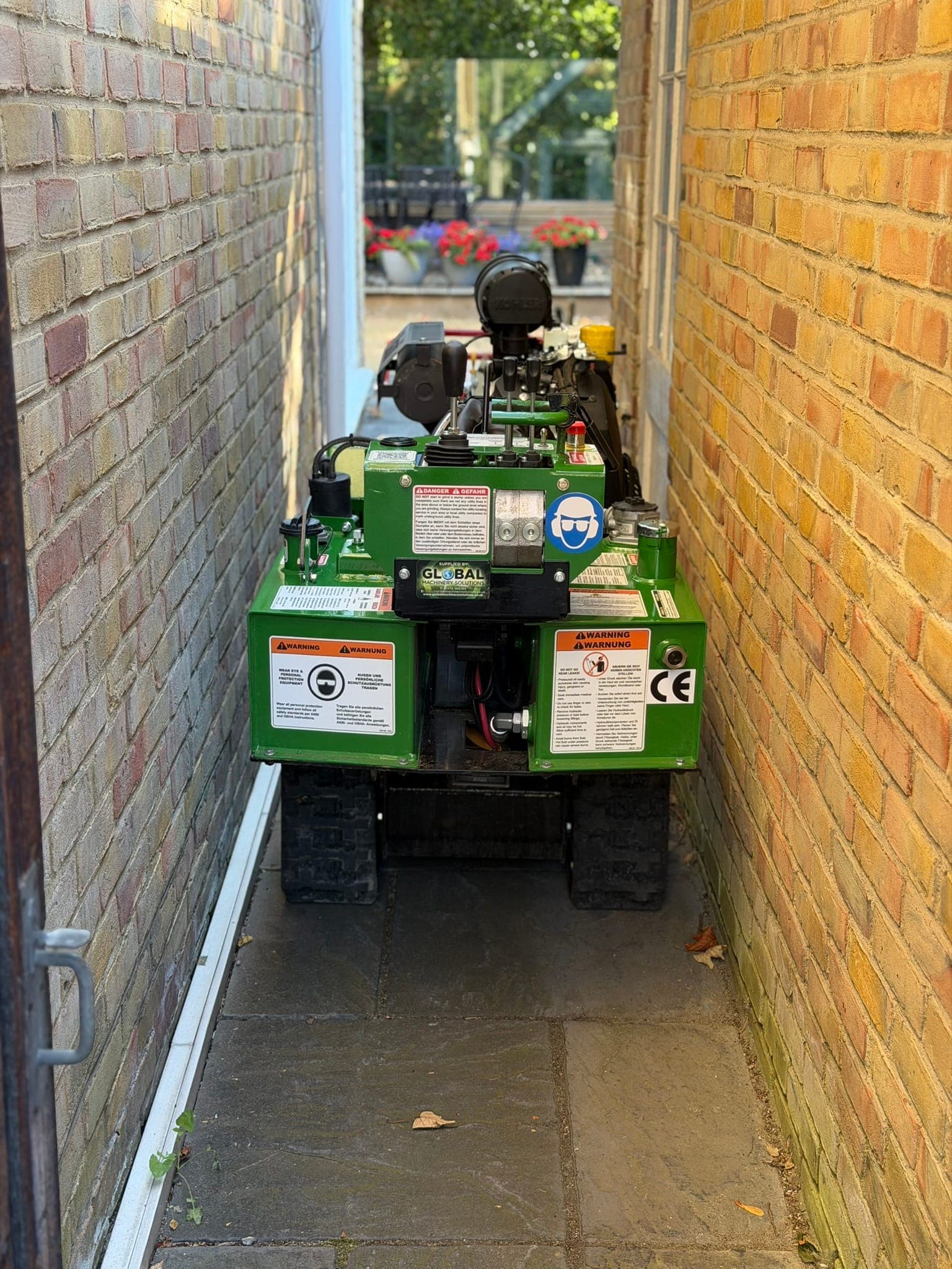 A green Bandit SG-40 stump grinder filling a narrow paved passage between two brick walls on its way to a rear garden