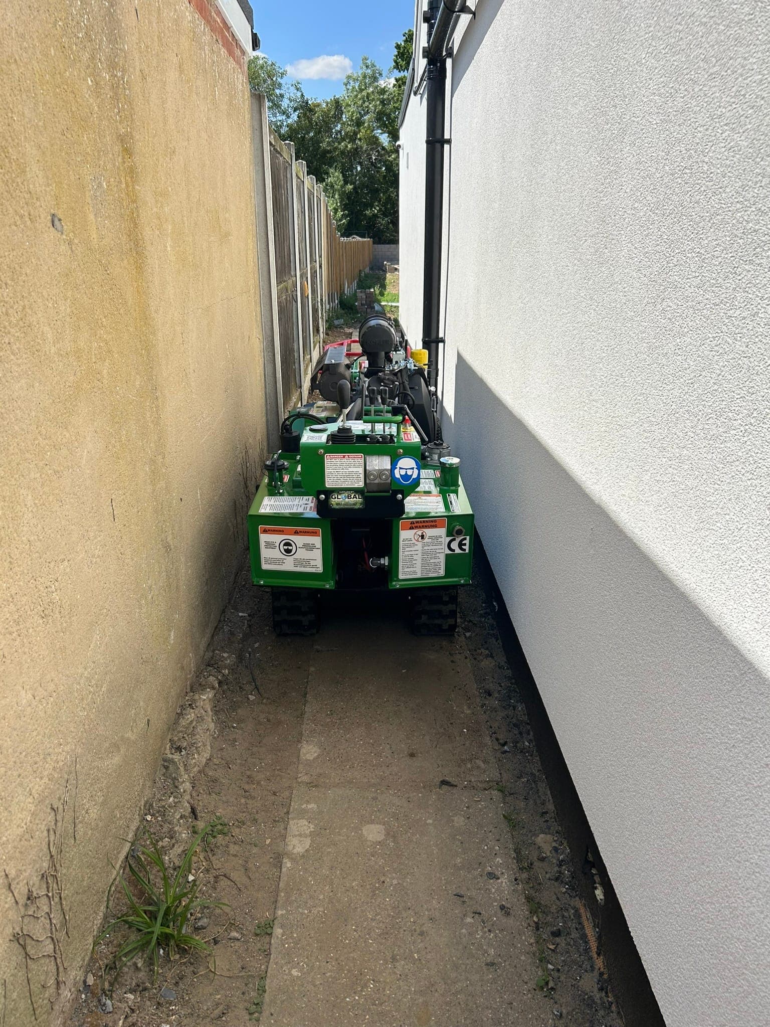 The Bandit SG-40 tracking along a narrow dirt side passage between a yellow rendered wall and a white rendered house, with a downpipe close to one side
