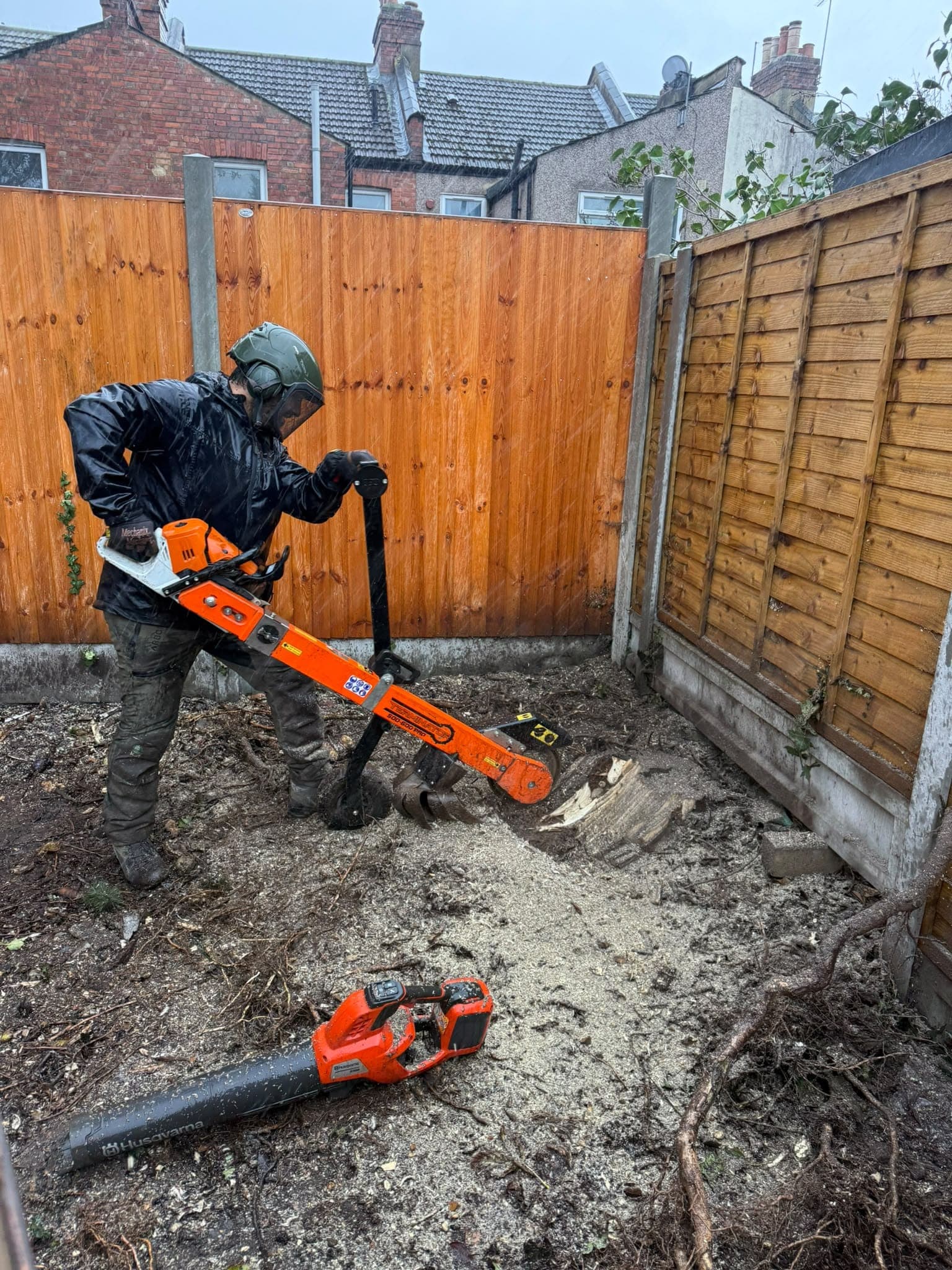 An operator in a helmet and visor grinding a stump with the Terminator attachment in the rain, in a small rear garden enclosed by close-board fences