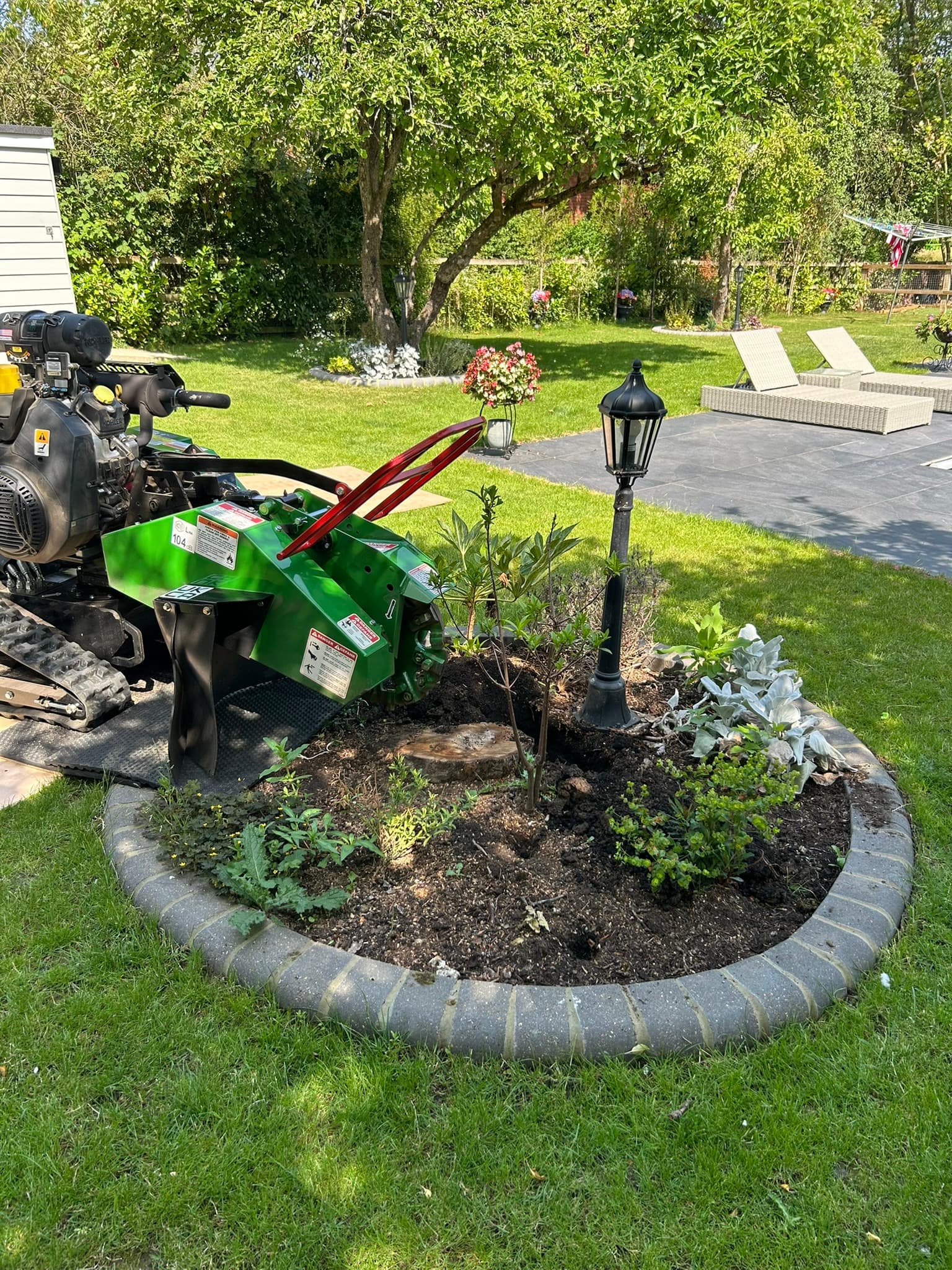 A tracked stump grinder working on a stump within a circular planted bed on a domestic lawn, with protective matting laid over the grass