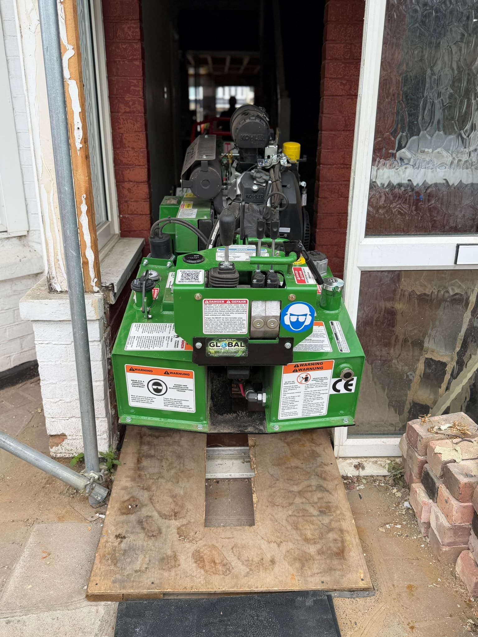 A green Bandit stump grinder at the front doorway of a house in Ilford, on plywood boards over the threshold, with the door frame removed for clearance