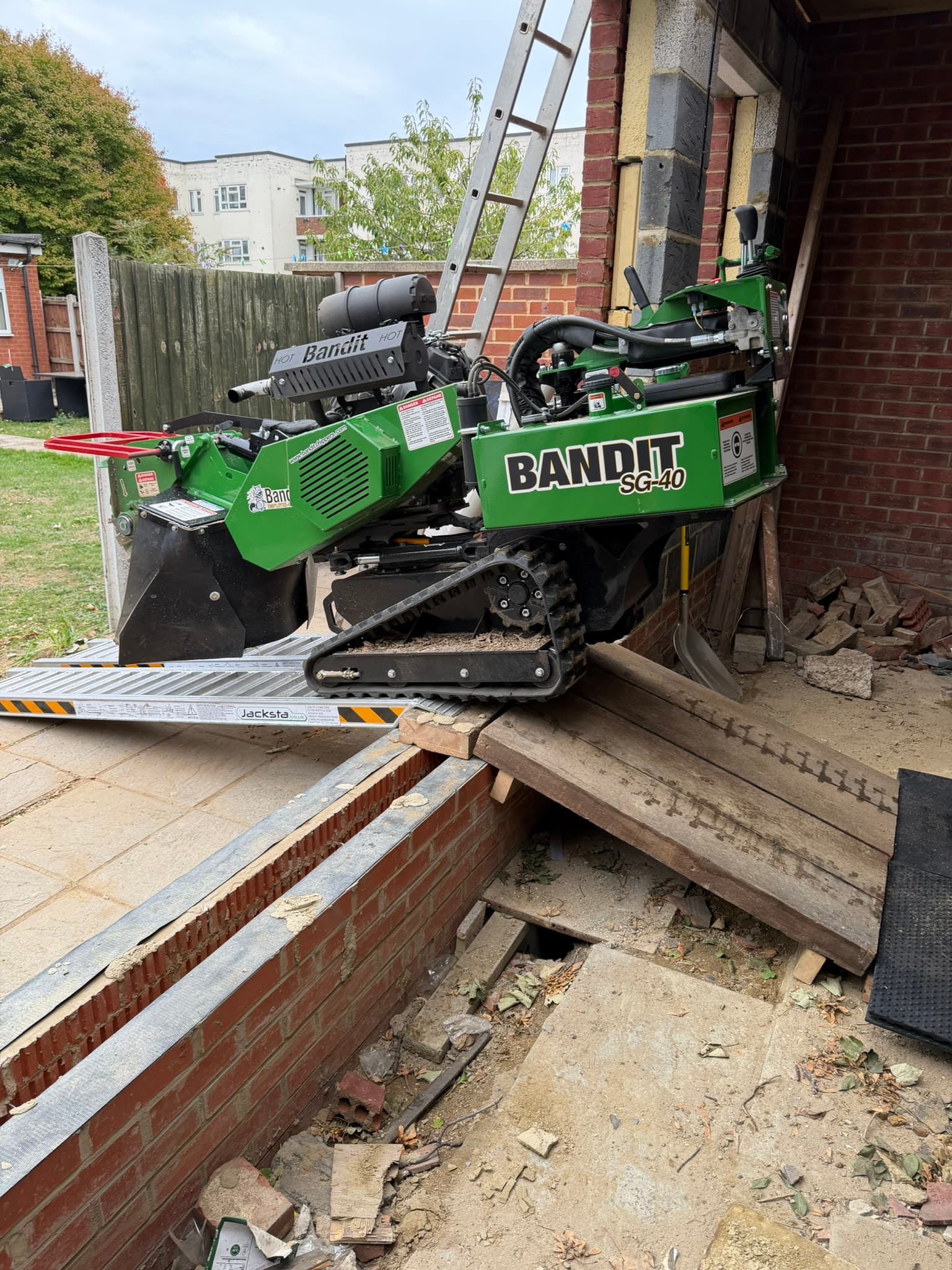 The Bandit SG-40 crossing a low brick wall on aluminium loading ramps and heavy timber sleepers to reach a rear garden in Ilford