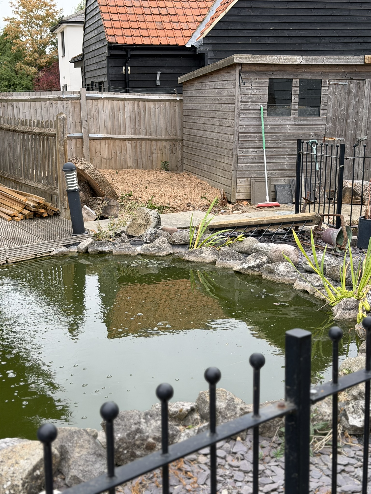 A wider view of the Brentwood garden after grinding, with fresh wood chippings against the fence beside a pond and outbuildings